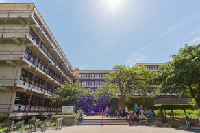 Chemiegebäude Chemiegebäude: Ein großes, breites, graues Gebäude mit Fensterfronten und Balkonen vor den Fenstern, die mit roten Metallstangen abgegrenzt werden. In der Mitte eine Treppe, auf der Menschen sitzen und links vor der Treppe Fahrräder in Fahrradständern.